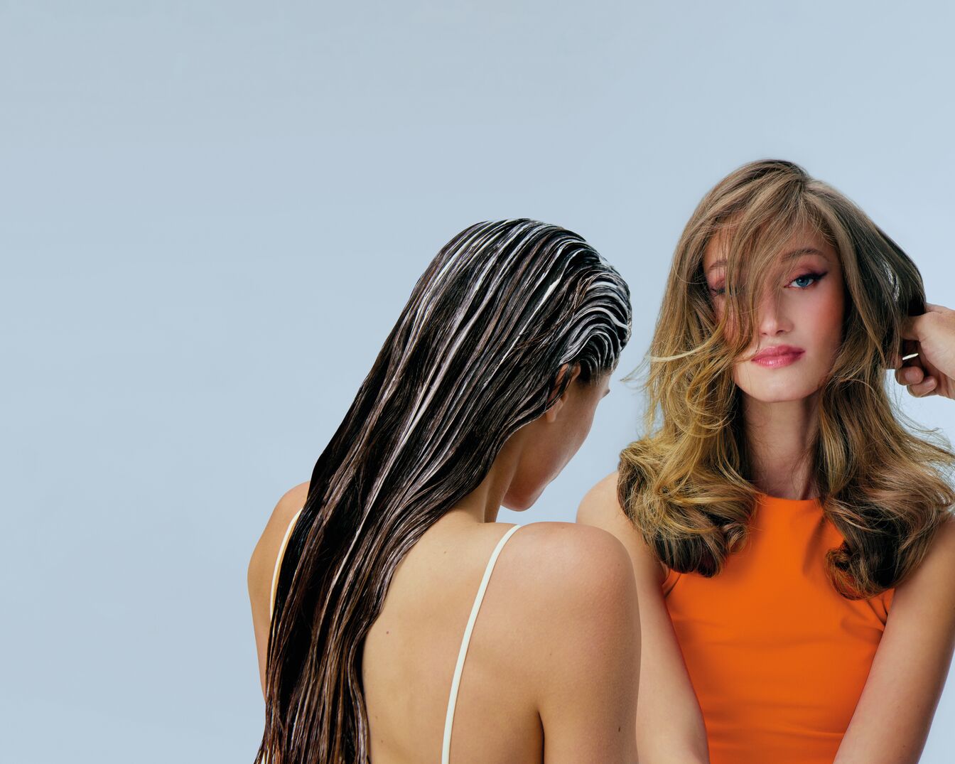 Sieviete ar tum&scaron;iem, mitriem matiem, kuriem uzklāts gai&scaron;s kondicionieris, stāv blakus sievietei ar blondiem, lokainiem matiem, ģērbtai oranžas krāsas apģērba aug&scaron;daļā.